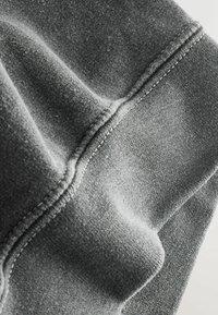 Pelēka auduma gabals ar mīkstu tekstūru un dažādām nokrāsām. Redzama šūšanas detaļa malās, kas izceļ gludu virsmu un krokas.