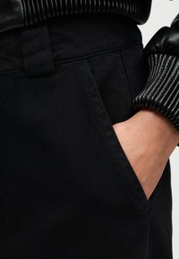 Χέρι στην τσέπη μαύρου παντελονιού, φοράει μαύρο ριμπ δερμάτινο μπουφάν με μανσέτα ορατή σε ανοιχτό φόντο.