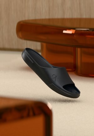 Sandale noire à bout ouvert avec logo "ck" flottant au-dessus d'un tapis beige dans une pièce avec des meubles translucides marron.