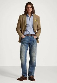 Mlad človek v svetlo rjavem blazerju, svetlo modri srajci, uničenih kavbojkah, rjavem pasu in rjavih čevljih, s rokami v žepih, ki stoji pred enostavno podlago.