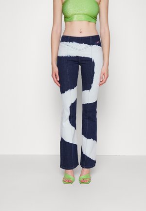 Femme portant un crop top vert, un jean évasé tie-dye bleu foncé et blanc, avec des talons verts à brides et bouts ouverts, debout devant un fond blanc.