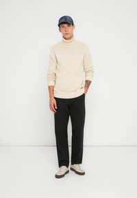 Beige turtleneck sweater, sorte bukser og hvide sneakers med brune såler. Båret med en blå kasket. Simpel design, tætsiddende form, glat tekstur.