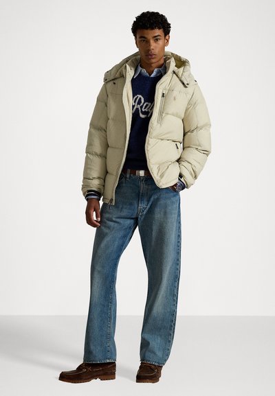 Beige puffer jacket with a hood, featuring a front zipper and side pockets. Paired with a navy sweater, light blue shirt, and blue jeans.