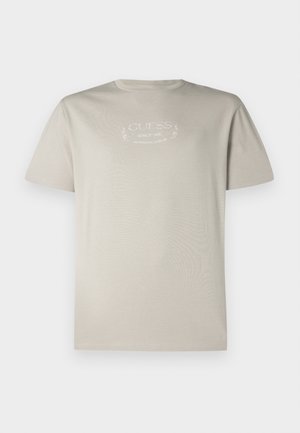 Camiseta de manga corta beige con cuello redondo y texto impreso en blanco que dice "GUESS Since 1981 Authentic Design" en el pecho.