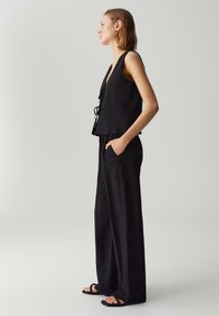 Zwarte mouwloze top met een V-hals en verstelbare strikjes, gecombineerd met losse zwarte broek met zijzakken en wijde pijpen.