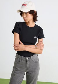 Camiseta ajustada negra de manga corta, con un pequeño logo blanco. Combinada con jeans ajustados de talle alto grises y una gorra beige con texto rojo.