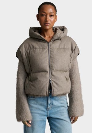Donna che indossa un gilet imbottito senza maniche color taupe con cappuccio sopra a una maglia pelosa a maniche lunghe e jeans azzurri chiari su sfondo neutro.