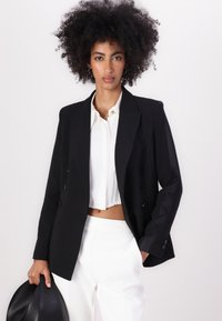 Blazer preto de dupla abotoadura com lapelas de pico, em tecido texturizado, apresentando dois bolsos frontais, combinado com uma blusa branca cropped e calças ajustadas.
