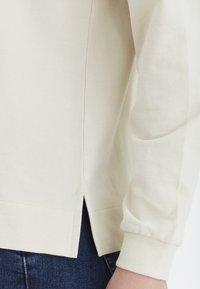 Camisa de manga larga beige claro con una tela ligeramente texturizada, presenta aberturas laterales y puños ajustados. Diseño minimalista, sin patrones ni acentos.