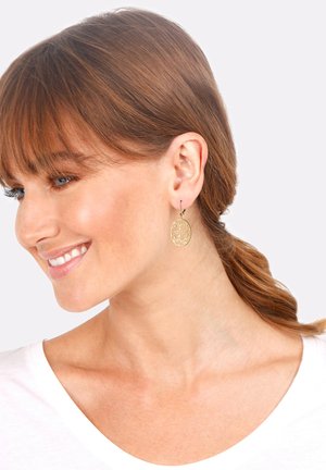 Boucles d'oreilles ovales dorées avec un design découpé complexe suspendues à un crochet simple. Présentées sur une personne aux cheveux mi-longs.