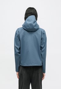 Μπλε αδιάβροχο μπουφάν με κουκούλα, με μινιμαλιστικό σχέδιο, ομαλή υφή και μακριά μανίκια. Πλάγια όψη από πίσω, φορεμένο πάνω από σκούρο παντελόνι.