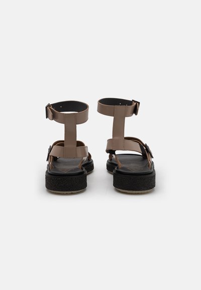 Brown faux leather sandals with a chunky black sole, adjustable ankle straps, and simple buckle details; open-toed and flat design.