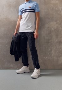 Paita polo vaaleansinisenä ja valkoisena, jossa on tummansiniset raidat, yhdistettynä mustiin cargo-housuihin ja valkoisiin urheilukenkiin. Pitää mustaa takkia kädessään.