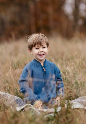 Sininen fleece-takki, jossa etukejyke ja ruskea nahkainen vetoketjun vedin, päällä lapsi istuu korkeassa ruohikossa. Taustalla näkyy syksyn lehtivihreitä.