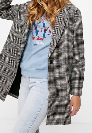 Femme portant un sweat-shirt New York bleu clair, un manteau à carreaux noir et blanc, et un jean bleu clair, posant avec un bras levé.