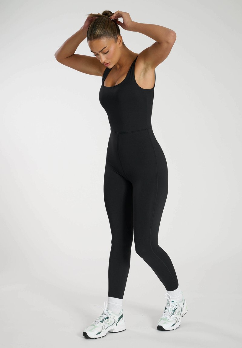 Zwarte aangesloten jumpsuit met brede bandjes, hoge halslijn en naadloos ontwerp. Kenmerkt zich door zachte stof en taps toelopende broekspijpen, gecombineerd met sportschoenen.