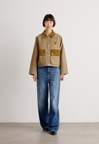 Bruine colbert met twee voorzakken, beige stof en gouden hardware; gecombineerd met losse blauwe jeans en zwarte schoenen.