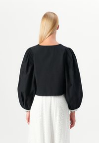 Blouse noire courte avec des manches longues bouffantes, dotée d'un col rond et d'une texture lisse, associée à une jupe blanche texturée.