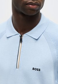Gros plan d'une personne portant un polo bleu clair à fermeture éclair avec le logo "BOSS" brodé sur la poitrine.