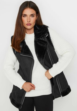 Schwarze Kunstlederweste mit einem weichen, schwarzen Fellkragen. Verfügt über einen diagonalen Reißverschluss und Seitentaschen. Getragen über einem weißen Strickpullover.