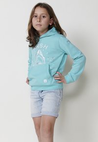 Smaragdzaļš kapuce ar haizivju grafiku un priekšējo kabatu, apvienots ar uzvilktiem gaiši ziliem džinsu šortiem. Mīksts audums, ikdienišķs dizains.