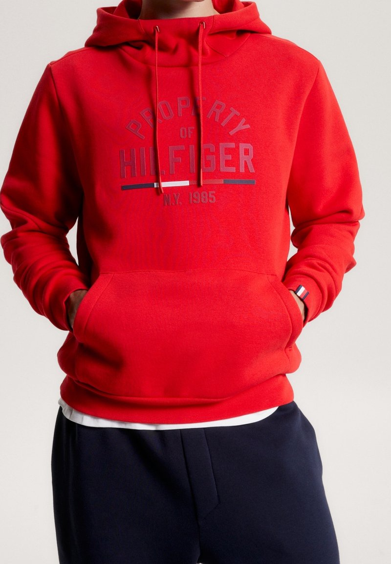 Felpa rossa con tasca a marsupio e cappuccio con coulisse. Presenta un logo centrale grigio con accenti blu e bianchi. Tessuto dalla texture liscia.