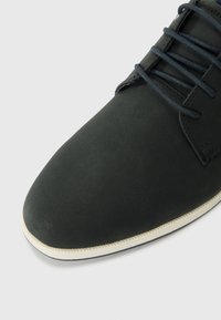 Black suede shoe with a rounded toe, lace-up design, and a contrasting white sole. Notable smooth texture and stitching detail along the edge.