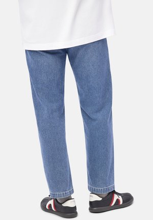 Person som bär blå rakbenade jeans, grå strumpor och svarta sneakers med röda och vita detaljer, stående framför en vit bakgrund.