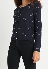Granatowy kardigan z miękkiego materiału, z okrągłym dekoltem i wzorami złotych półksiężyców, z białymi guzikami i ściągaczami na mankietach.