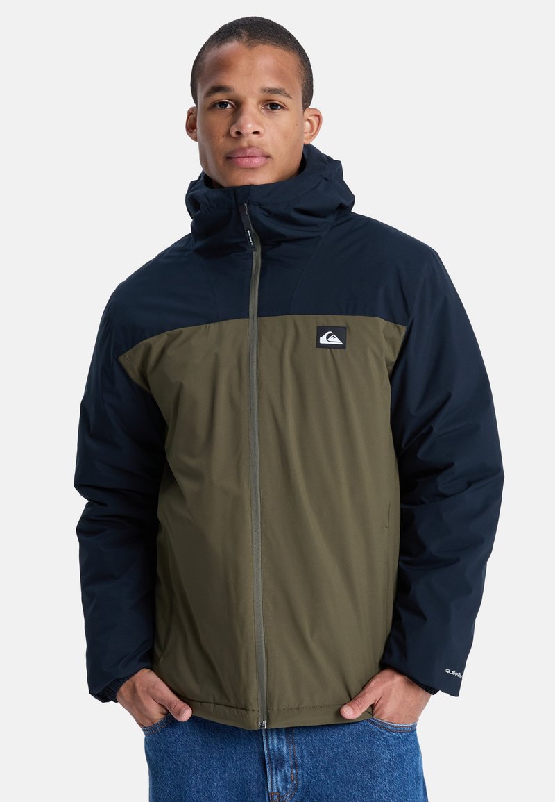Ung mand iført en tofarvet sort og olivenjakke med hætte, der står med hænderne i lommerne på blå jeans mod en ensfarvet baggrund.