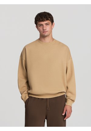 Felpa oversized beige con scollatura rotonda e polsini a costine, abbinata a pantaloni jogger marrone scuro. Texture del tessuto liscia, vestibilità rilassata.
