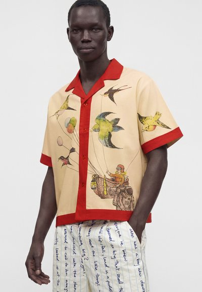 Homme portant une chemise beige à manches courtes avec col et poignets rouges, présentant des oiseaux et une personne dans un panier attaché à des ballons, assortie à un pantalon blanc avec un texte bleu.