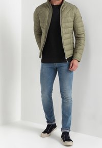 Olivgrüne Pufferjacke mit hohem Kragen, Reißverschluss, kombiniert mit einem schwarzen Pullover, hellblauen Jeans und dunklen Sneakern mit beigen Sohlen.