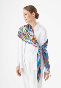 Écharpe en soie avec un motif floral dans des teintes de bleu, rose et orange, nouée élégamment autour des épaules sur une chemise blanche à boutons.