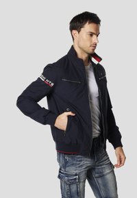 Námořnicky modrá bomber bunda se zipovými předními kapsami, žebrovanými manžetami a límcem s červenou podšívkou. Bílé logo „HTG“ na levém rukávu.