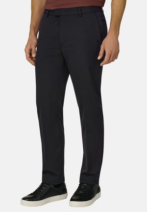 Mies, joka käyttää mustia slim fit -mallisia juhlahousuja etutaskuilla ja mustia matalia lenkkareita valkoisilla pohjilla, seisoo yksiväristä taustaa vasten.