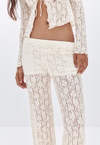 Spitzen-Crop-Jacke und passende hochtaillierte Hose in Creme mit floralen Mustern. Der Stoff ist transparent mit einer zarten, strukturierten Oberfläche.