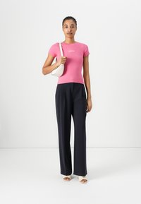 Figurbetontes pinkes Ripp-T-Shirt mit kurzen Ärmeln und kleinem Logo, kombiniert mit hochgeschnittenen navyblauen Hosen und einer weißen Umhängetasche.