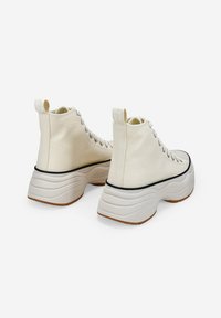 Zapatillas altas de color crema con parte superior de lona texturizada, suela de goma gruesa y acentos en negro a lo largo del borde de la suela.