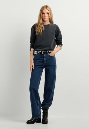 Giovane donna con lunghi capelli biondi che indossa una felpa nera, una camicia a righe, jeans blu a vita alta e stivaletti neri, in piedi con una mano in tasca.