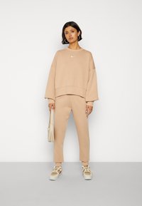 Sweatshirt oversized em algodão bege com logótipo branco, combinado com calças de treino ajustadas a condizer. O modelo segura uma pequena mala de creme e usa ténis.