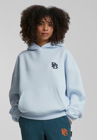 Felpa oversized azzurra chiara in misto cotone con tasca frontale. Presenta un logo ricamato blu scuro sul petto. Maniche a costine.
