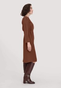 Braunes langärmliges Kleid mit gerafftem Tailleneinsatz, seitlichen Schlitzen und glatter Textur, kombiniert mit knielangen braunen Stiefeln, seitlich betrachtet.