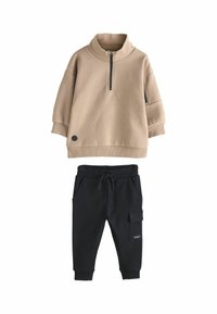 Beige Zip-Up-Sweatshirt mit hohem Kragen und schwarzen Akzenten, kombiniert mit schwarzen Jogginghosen mit Taschen und einem elastischen Bund mit Kordelzug.