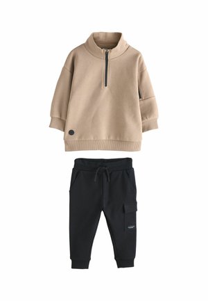 Beige Zip-Up-Sweatshirt mit hohem Kragen und schwarzen Akzenten, kombiniert mit schwarzen Jogginghosen mit Taschen und einem elastischen Bund mit Kordelzug.
