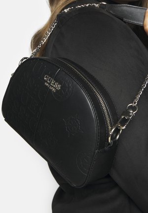 Sac bandoulière noir Guess avec motif logo embossé et chaîne argentée porté sur l'épaule contre un vêtement noir.