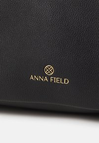 Sac en cuir noir texturé arborant un logo doré "ANNA FIELD" avec un accent rond, positionné dans la section inférieure.