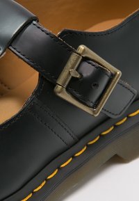 Chaussure en cuir noire avec une boucle en métal, des surpiqûres jaunes le long de la semelle, et une texture lisse avec un design arrondi.