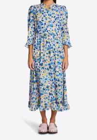 Kwiatowa sukienka midi z niebieskimi, żółtymi i różowymi wzorami, okrągłym dekoltem, bufkami na rękawach i falbaną u dołu. W zestawie z jasnoróżowymi chodakami.