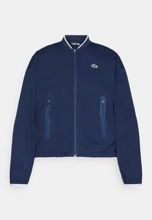 Chaqueta bomber azul marino con cremallera frontal, dos bolsillos laterales con cremallera y puños elásticos. Presenta un pequeño logo verde de Lacoste en el pecho.
